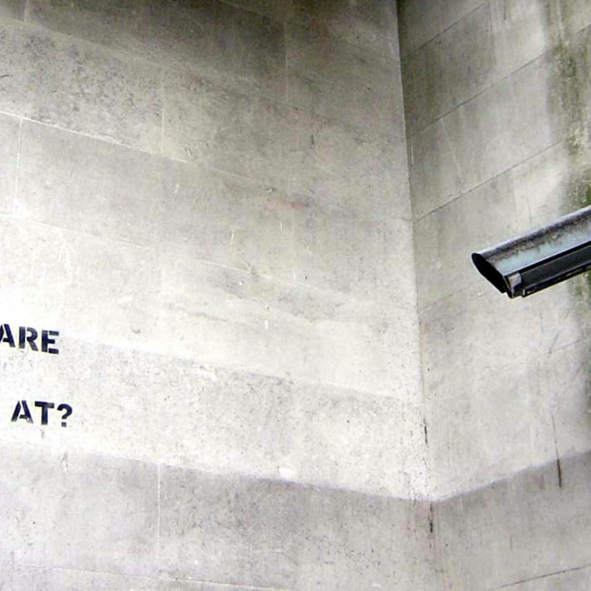 Photograph of a CCTV camera pointing at the stencilled words "What are you looking at?"