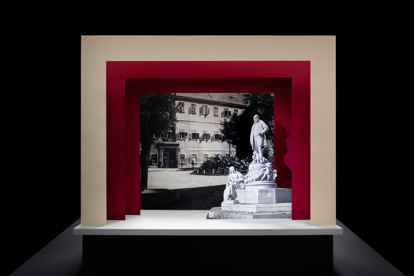 Photograph of a simple theatre stage set, made out of card. The background surrounding the stage is black. The stage floor is white and the framing of the stage is made out of 3 square edged arches, each one smaller than the other, receding backwards. The first arch is cream coloured and the other two are a red. On the stage is a small cut out photograph of a grand statue man with a beard from the early 20th century. Behind him forming the backdrop is a black and white archive photograph of a the outside of a hospital building.