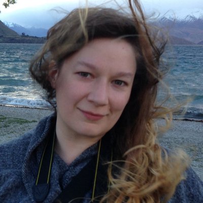 Head and shoulders photograph of a woman with long hair, blown around by the wind, with a grey pebble beach and the sea in the background.