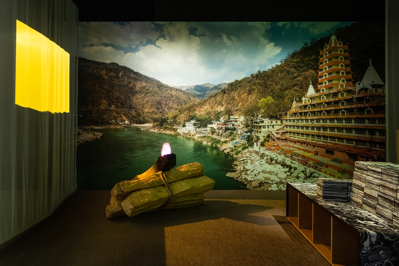 Photograph of a dark exhibition gallery space showing a constructed scene. On the far wall is a very large photographic print showing the sea, a rocky shoreline, a large temple like building and a mountain ranges. In front of this wall is a bungle of large cut wooden logs, lying on the floor on their side, which are tied around the middle. Above the rope is a purple flame like object. To the left of the logs is a white hanging curtain onto which a yellow rectangle is projected. To the far right is a wooden bench seat covered in a black and white patterned veneer.