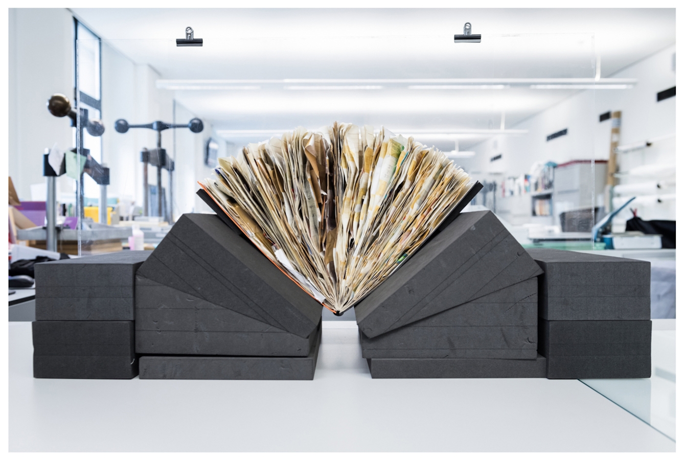 Photograph showing a spiral bound scrapbook resting on grey foam wedged book supports. The scrapbook is filled with material such that the pages are fanning out under the pressure The book is in a conservation studio, surrounded by various pieces of equipment.