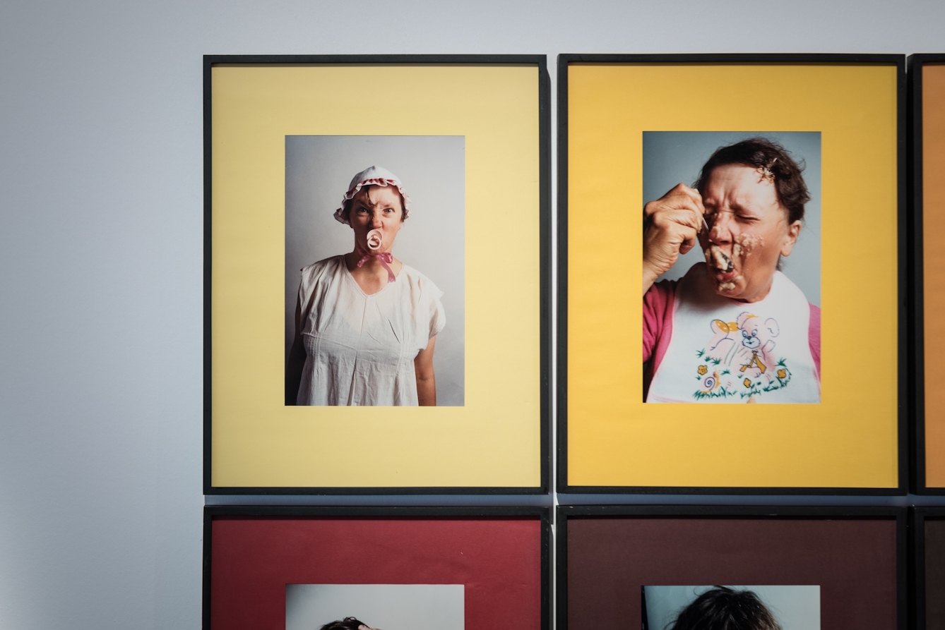 Photograph of two framed prints from a larger grid of images. The image on the left shows a photograph of a woman wearing a baby's bonnet and sucking a dummy, framed in a yellow mount. On the right is an image of a woman wearing a baby's bib, feeding herself with a spoon. Her face is covered in food. The image is framed in a yellow mount.
