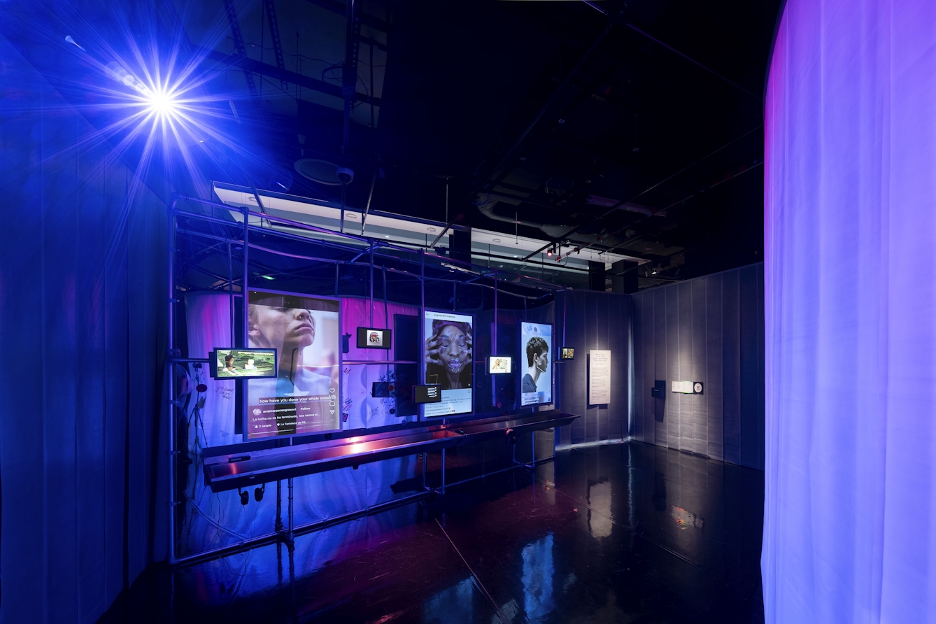 A art installation evoking a nightclub bathroom with images of faces projected on the wall above a long continuous sink in a darkened exhibition space with curtains hanging from the ceiling on either side and a very bright spot light shining  at the top left corner.