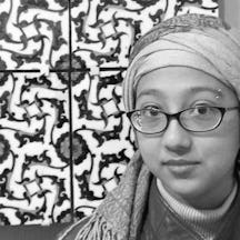 Black and white photograph of Shireen Hamza, a brown woman wearing a turban, glasses, and a jacket, standing in front of a floral ceramic design.