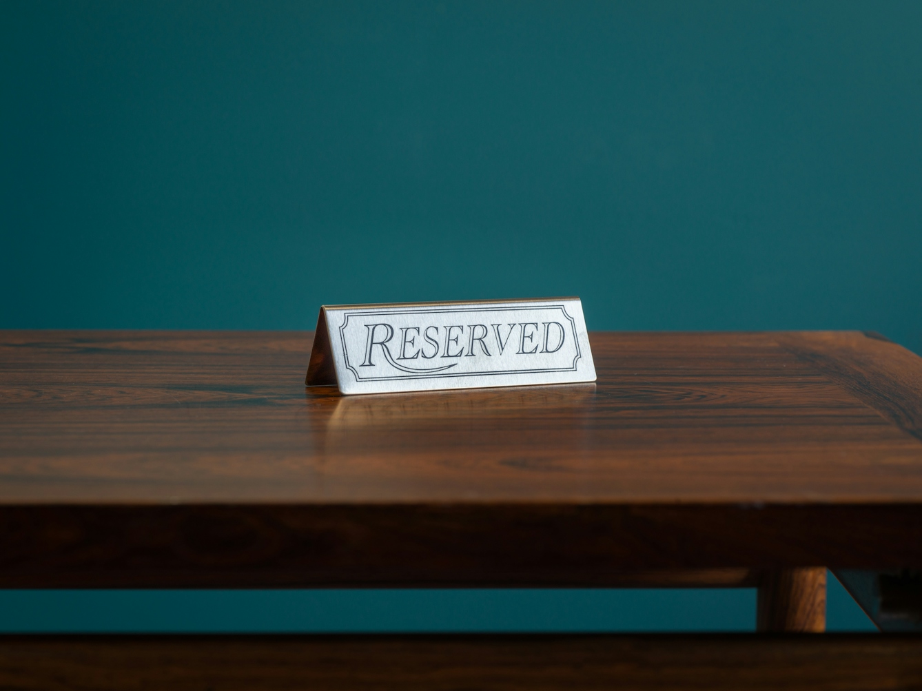 A photograph of a dark wood tabletop against a teal coloured background. On the table top is a small metal sign with the text ‘reserved’ etched on it.
