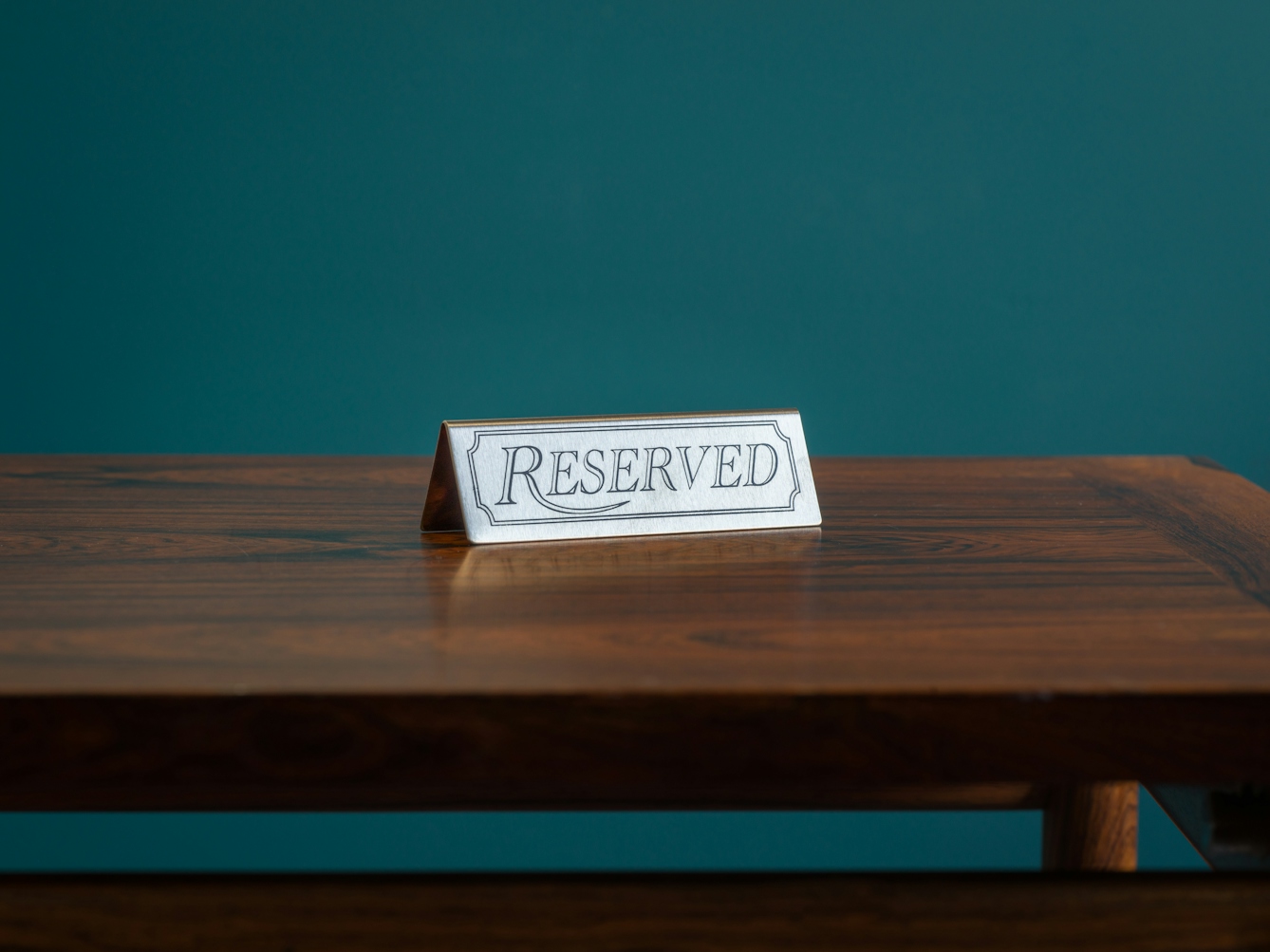 A photograph of a dark wood tabletop against a teal coloured background. On the table top is a small metal sign with the text ‘reserved’ etched on it.