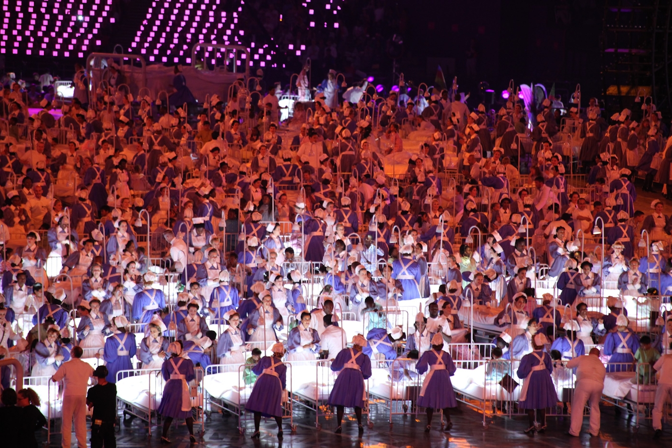 Photograph of many people dressed in historic nursing uniform of blue dresses and bonnets wheeling hospital beds containing children around as part of the 2012 Olympics Opening Ceremony. 