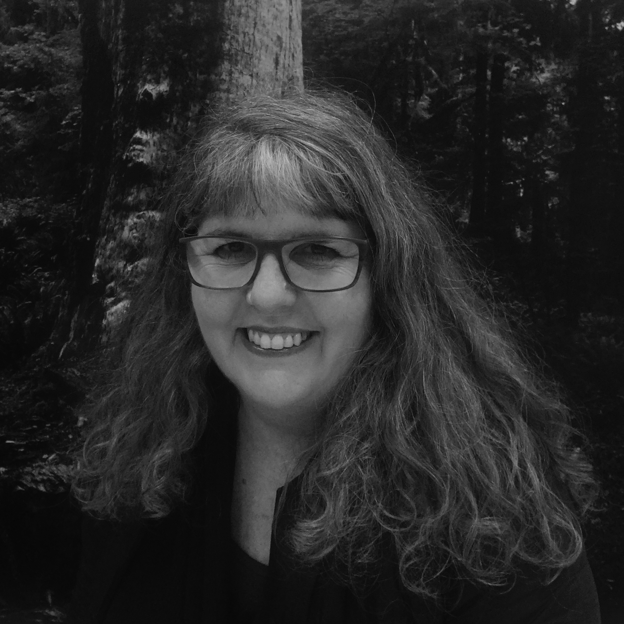 Black and white, head and shoulder photograph of a woman with long, curly hair and glasses wearing a jacket. She is standing in a forest area.
