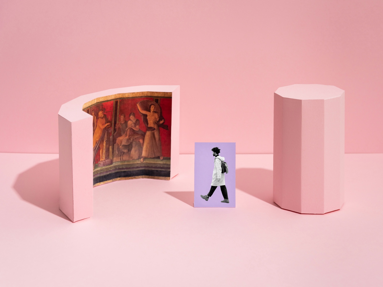 Photograph of a set built scene. The scene is made out of a light pink horizontal surface against a light pink vertical background with a thin horizon line. On the horizontal surface are two pink three dimensional shapes. The shape on the right is a tall thin shape with ten vertical sides. The shape on the left is semicircular with a Roman fresco visible on the inside curve. In the centre of the image is a rectangular flat purple card standing upright, on which is the black and white image of a man in full length wearing a backpack walking towards the fresco.