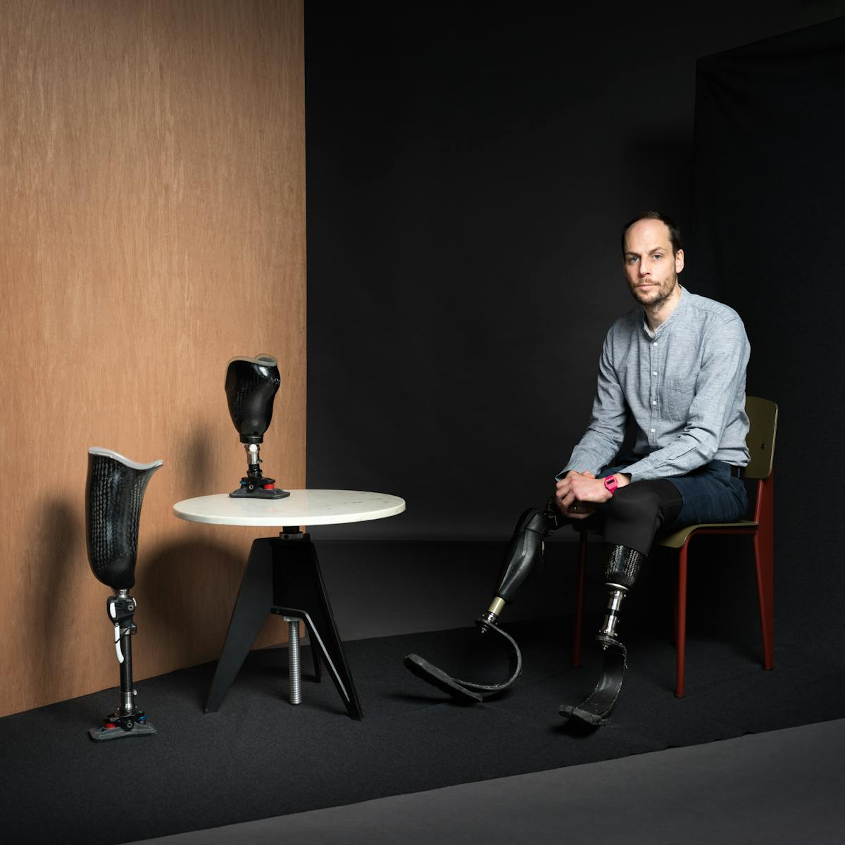 Portrait photograph of Harry Parker. He is wearing a grey shirt and jeans. He has short brown hair and a beard. Harry is sitting on a chair, his prosthetic limbs are resting on the ground. Next to his chair is a white table which has a prosthetic limb upon it. On the floor next to the table is another prosthetic limb standing upright and behind this, a wooden panel. 