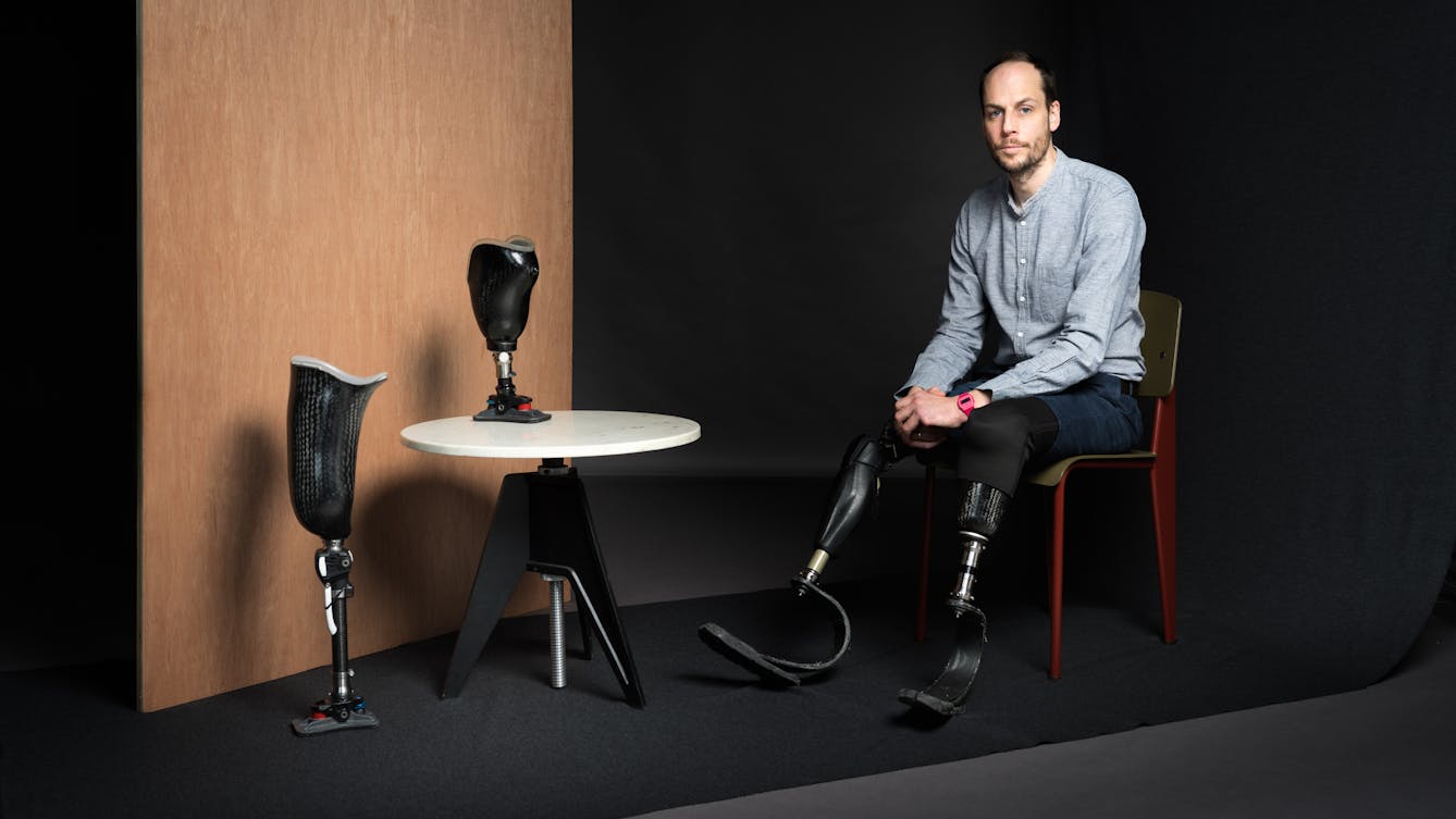 Portrait photograph of Harry Parker. He is wearing a grey shirt and jeans. He has short brown hair and a beard. Harry is sitting on a chair, his prosthetic limbs are resting on the ground. Next to his chair is a white table which has a prosthetic limb upon it. On the floor next to the table is another prosthetic limb standing upright and behind this, a wooden panel. 
