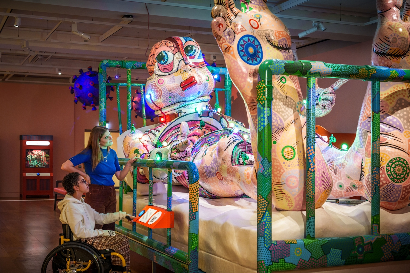 Photograph of two people looking at the giant sculpture of Jason lyng in a hospital bed.