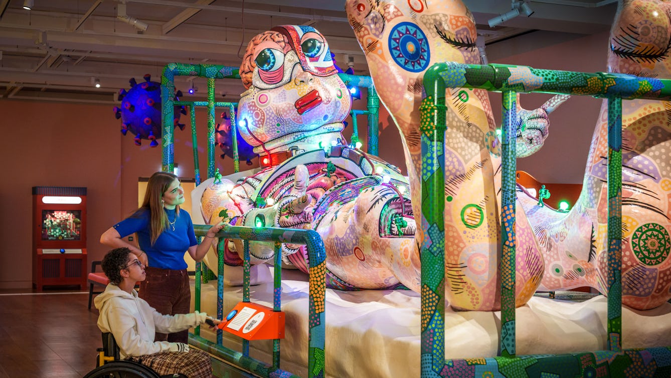 Photograph of two people looking at the giant sculpture of Jason lyng in a hospital bed.