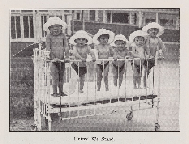 Black and white photo of six young children standing in a cot, wearing white sun hats.