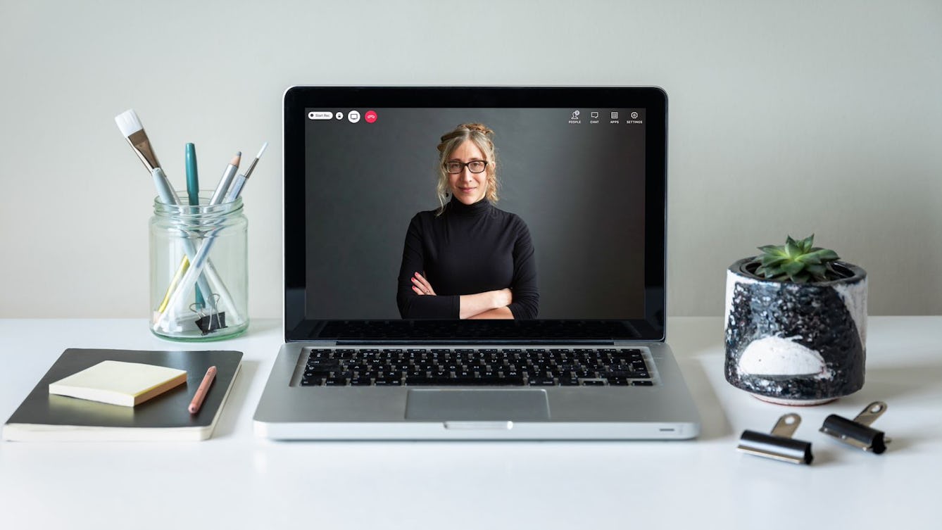 Photo of a laptop with a video chat image of a woman in a black polo neck. On the table are a potted plant, a glass jar with stationery inside and a notepad, post-it notes and a pencil.