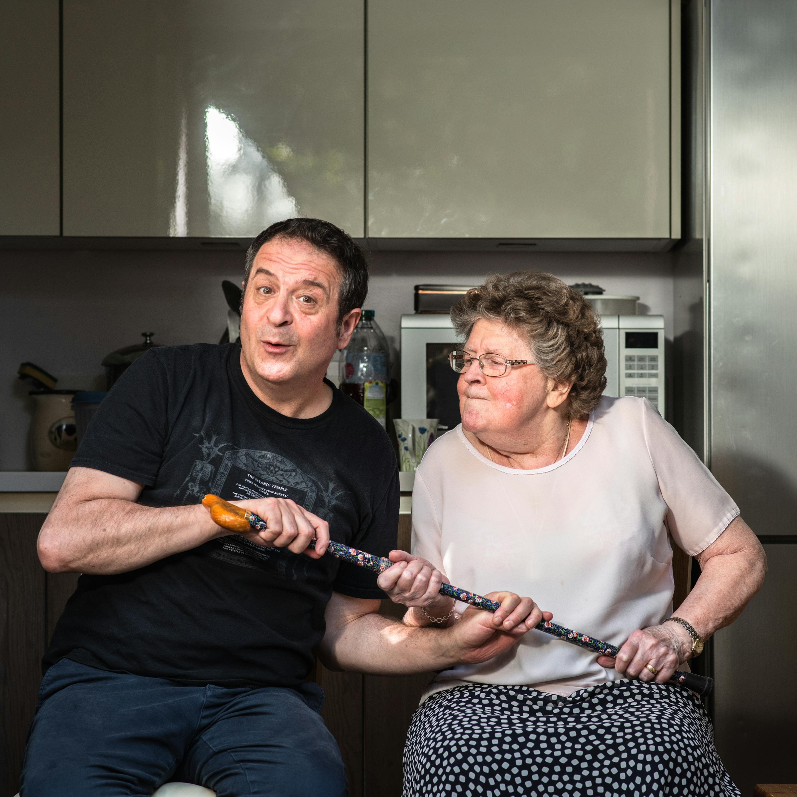 Photograph of a man and his mother sat side by side in a kitchen. The man wearing a black t-shirt and blue trousers is seated on the left has a cheeky look on his face, looking straight to camera. His mother on the right is wearing a pink blouse and blue patterned skirt and is looking towards him with pursed lips and a slightly annoyed and determined look on her face. She is thrusting her walking stick towards him with both hands, handle end first. Her son also has both his hands clasped to the stick. It looks as though they are both having a tussle over the stick. In the background are grey kitchen units, a microwave, the tap of the sink and a large silver fridge.