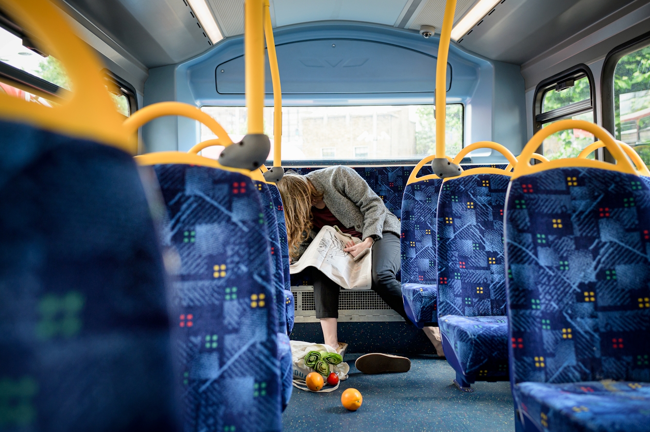 Photograph of a woman slumped on the back seat of the upper deck of a bus, a newspaper scrunched on her lap. On the floor in front of her, one of her shoes has come off and her bag of groceries has fallen over and a couple of oranges have escaped.