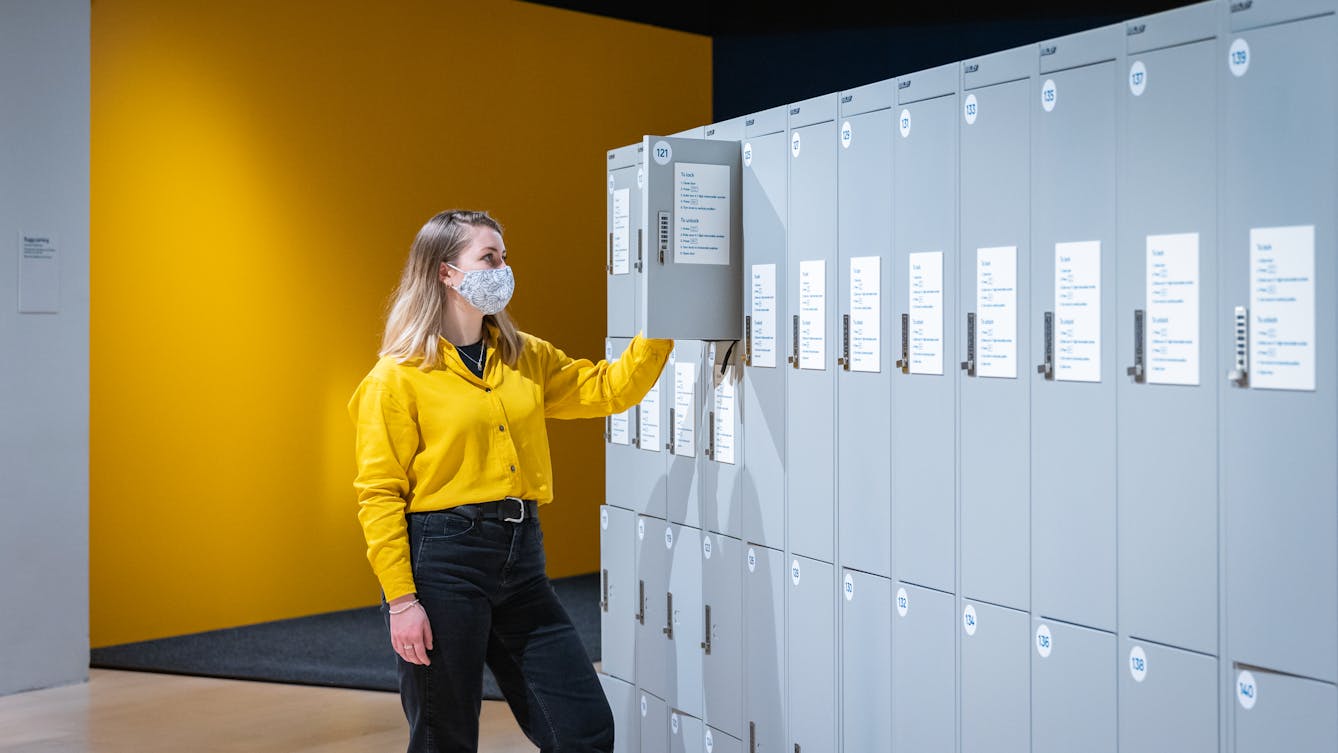 Photograph of a young woman wearing a face covering, a yellow shirt and black trousers, standing in front of a row of grey lockers. One of the locker doors is open and the woman is in the process of putting an item into the locker. Behind her is the yellow wall of the room.