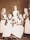 Photograph of five women, three standing and two seated in front of them. They wear Victorian clothing including caps, dark blouses, and white aprons.