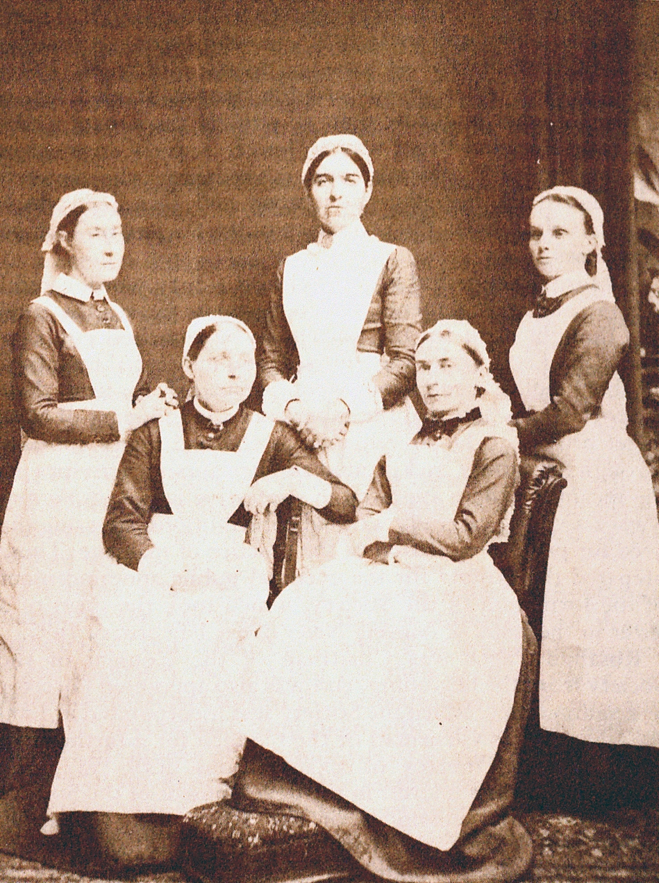 Photograph of five women, three standing and two seated in front of them. They wear Victorian clothing including caps, dark blouses, and white aprons.