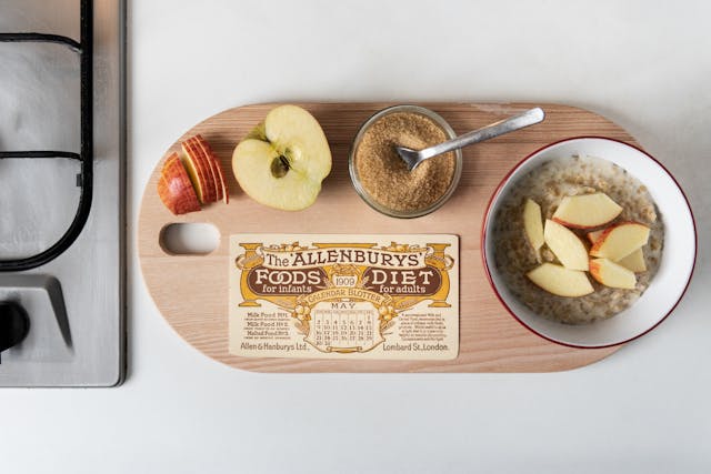 Photograph showing a wooden chopping board on a kitchen cabinet, next to a gas hob. On the chopping board, there is a sliced apple, a ramekin of sugar with a teaspoon in it and a bowl of porridge topped with apple slices. Alongside the food, there is a calendar blotter which reads 