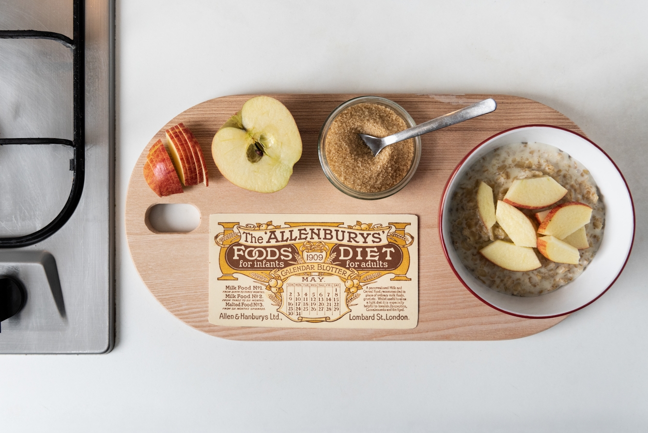 Photograph showing a wooden chopping board on a kitchen cabinet, next to a gas hob. On the chopping board, there is a sliced apple, a ramekin of sugar with a teaspoon in it and a bowl of porridge topped with apple slices. Alongside the food, there is a calendar blotter which reads 'The 'Allenburys' Food for infants, Diet for adults: 1909'. Below the text there is a small printed calendar. 