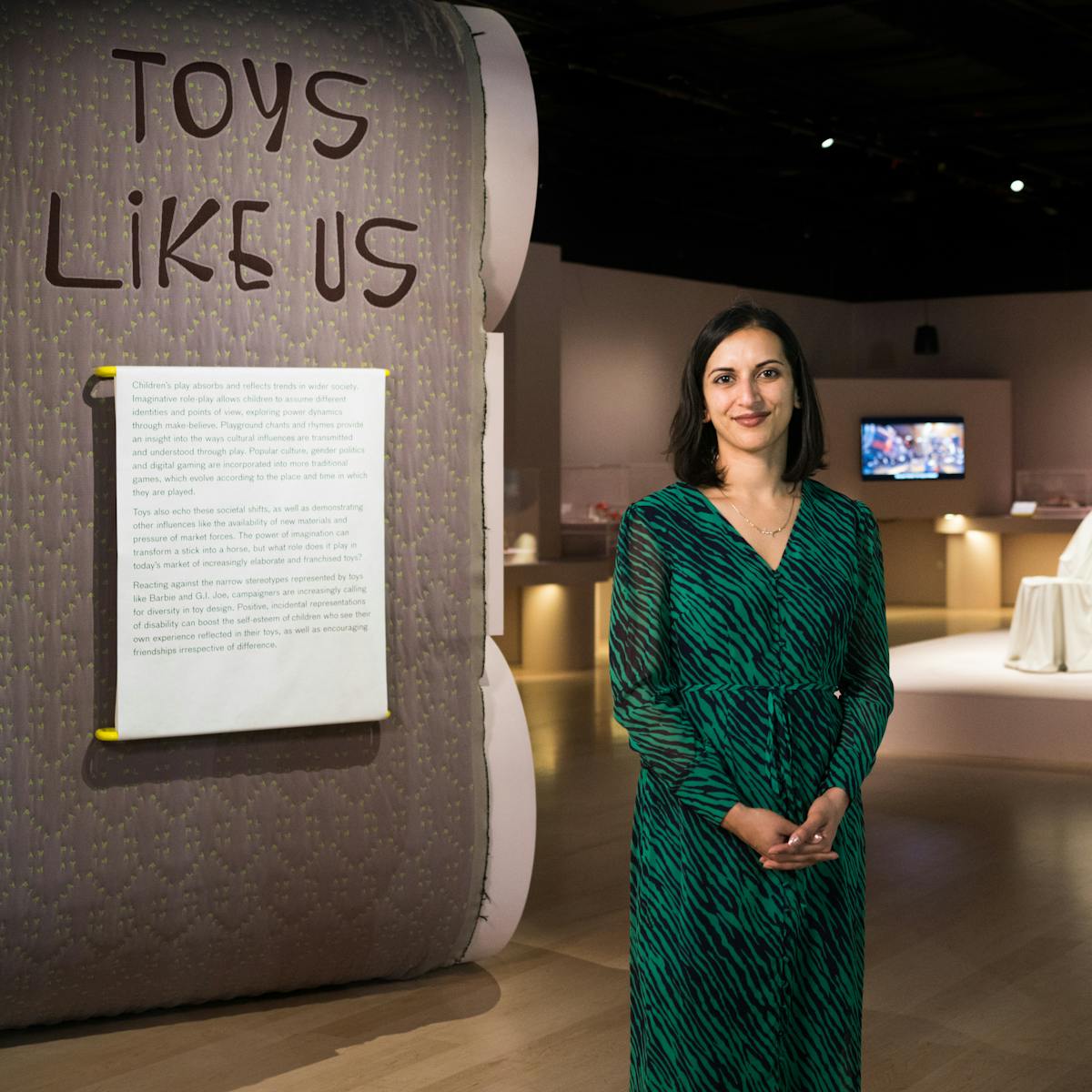 Photograph of a woman standing an exhibition gallery smiling to camera. To the left of her is an exhibition text panel with the title 'Toys Like Us' written above it. In the distance behind her to the right are exhibition exhibits and a tv screen.