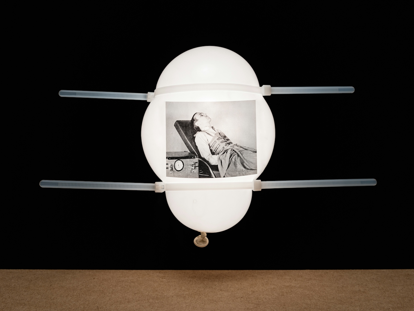 Photograph of a white inflated balloon against a black background, floating vertically above a wooden tabletop horizon line. The balloon looks like it is illuminated from within. On the side of the balloon is a rectangular, monochrome archive film still. The still shows a man lying propped upright on a bed with a contraption made of straps wrapped around his chest. To his right is a mechanical box which is connected to the chest contraption.  The two work together as an artificial ventilator. The balloon has 2 white cable ties around its surface which are deforming the balloon's shape and  echoing the contraption shown in the film still.