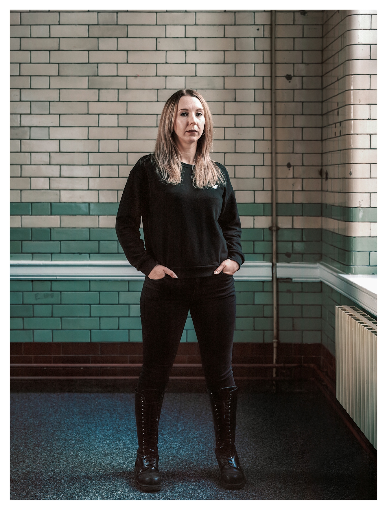 Photographic full length portrait of a woman standing looking to camera with her hands resting in the pockets of her black trousers. She is wearing a black long sleeved top and knee high lace up black boots. She is lit by a soft light from a large window just out of frame to the right. Behind her are the glossy tiled walls of a Victoria period building. The tiles are beige and green and are arranged in a horizontal pattern.