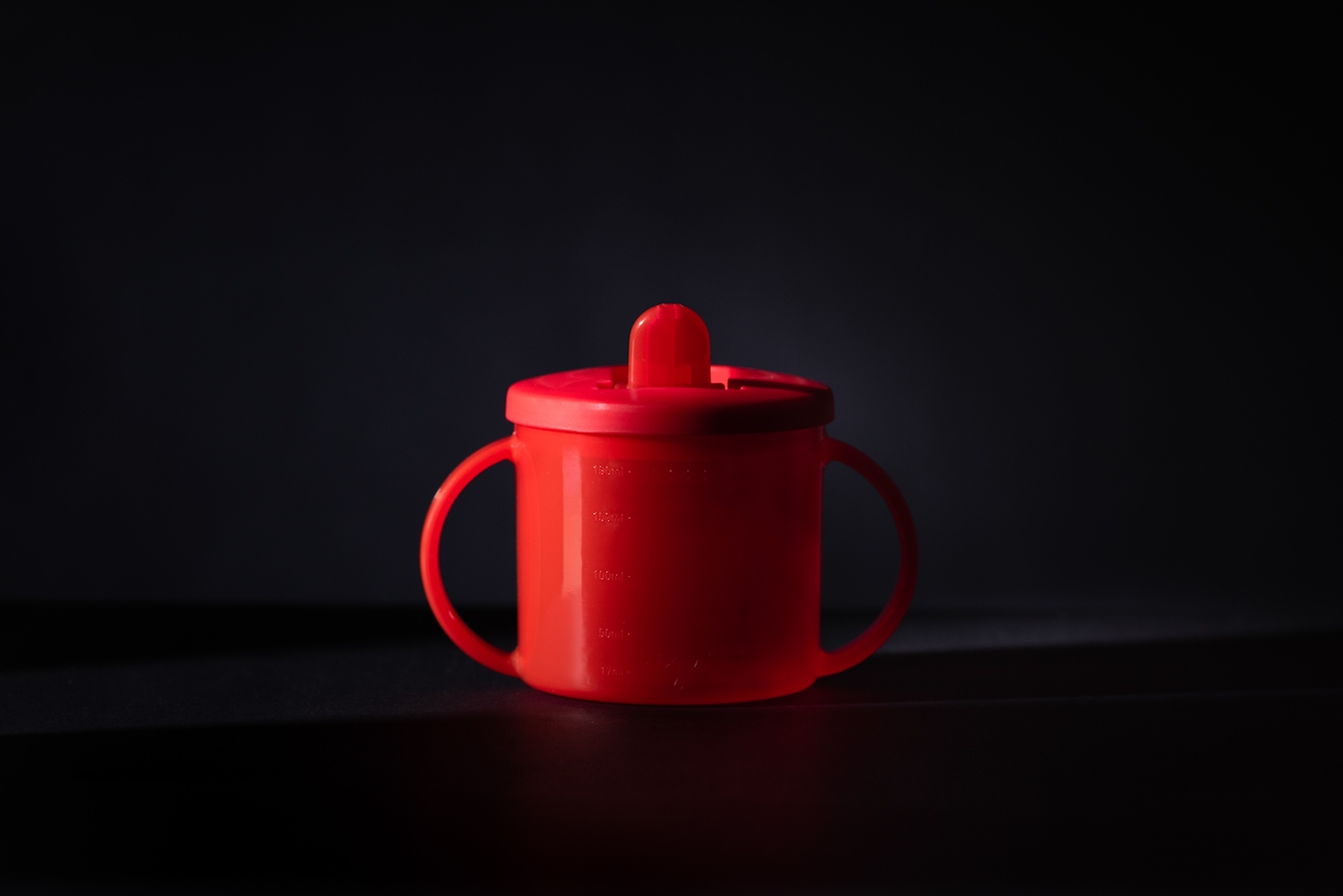A photograph of a toddler sip cup against a black background. The cup is red with two handles, a spout and appears so that one half is in dark shadow and silhouette.