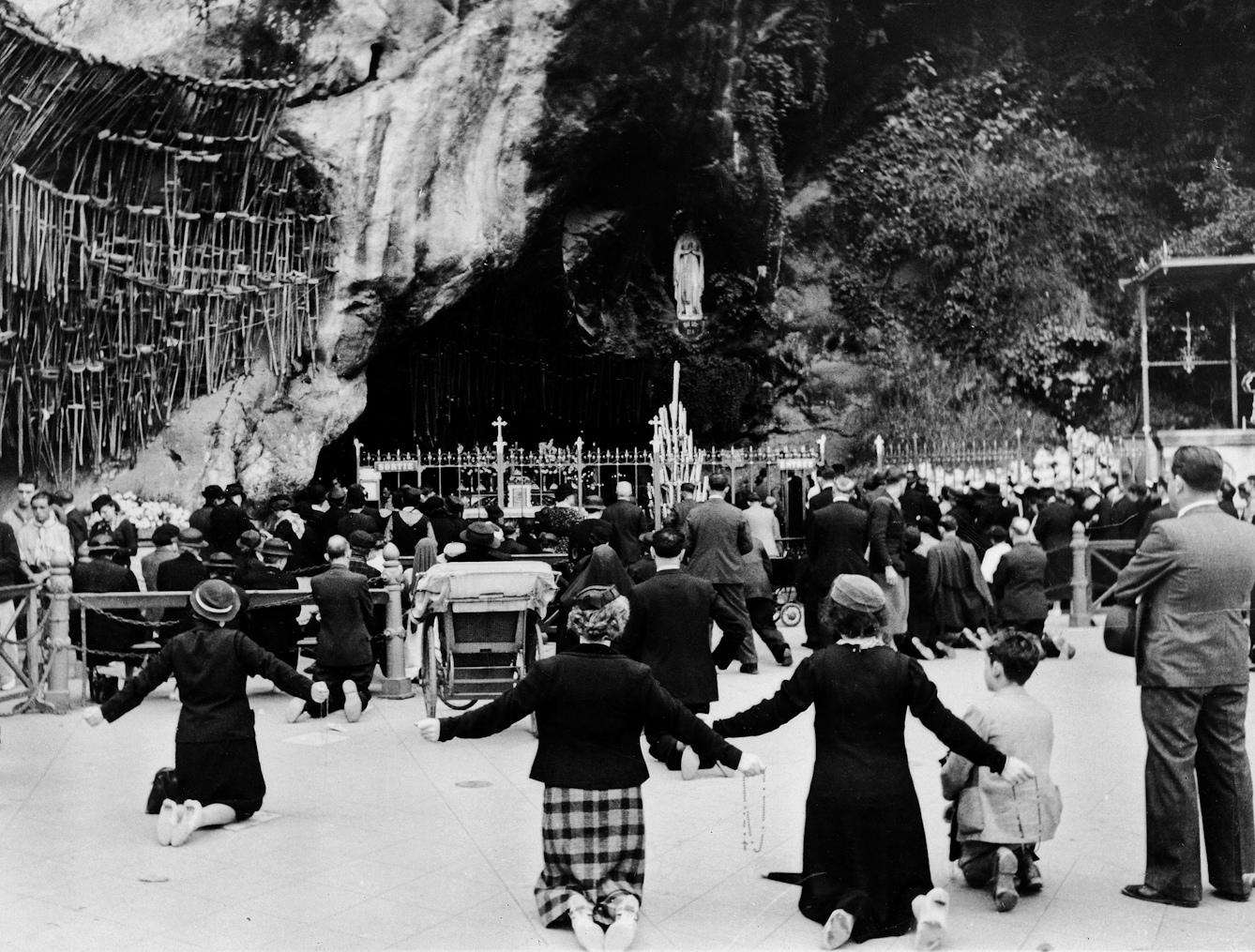 Black and white photograph of people kneeling at a shrine in Lourdes. Beside the worshippers, crutches can be seen hanging up.