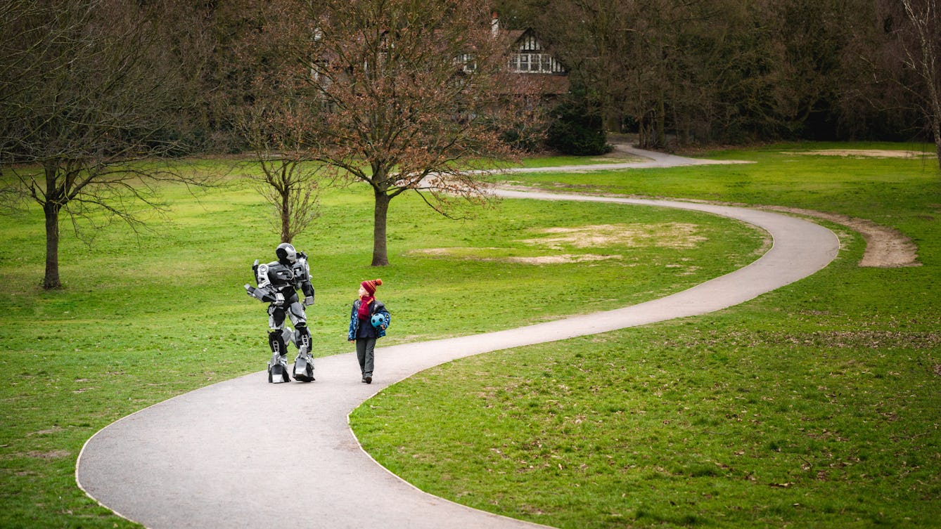 Photograph of a large robot walking and talking with a young boy in school uniform who's holding a football, down a winding path in the park.