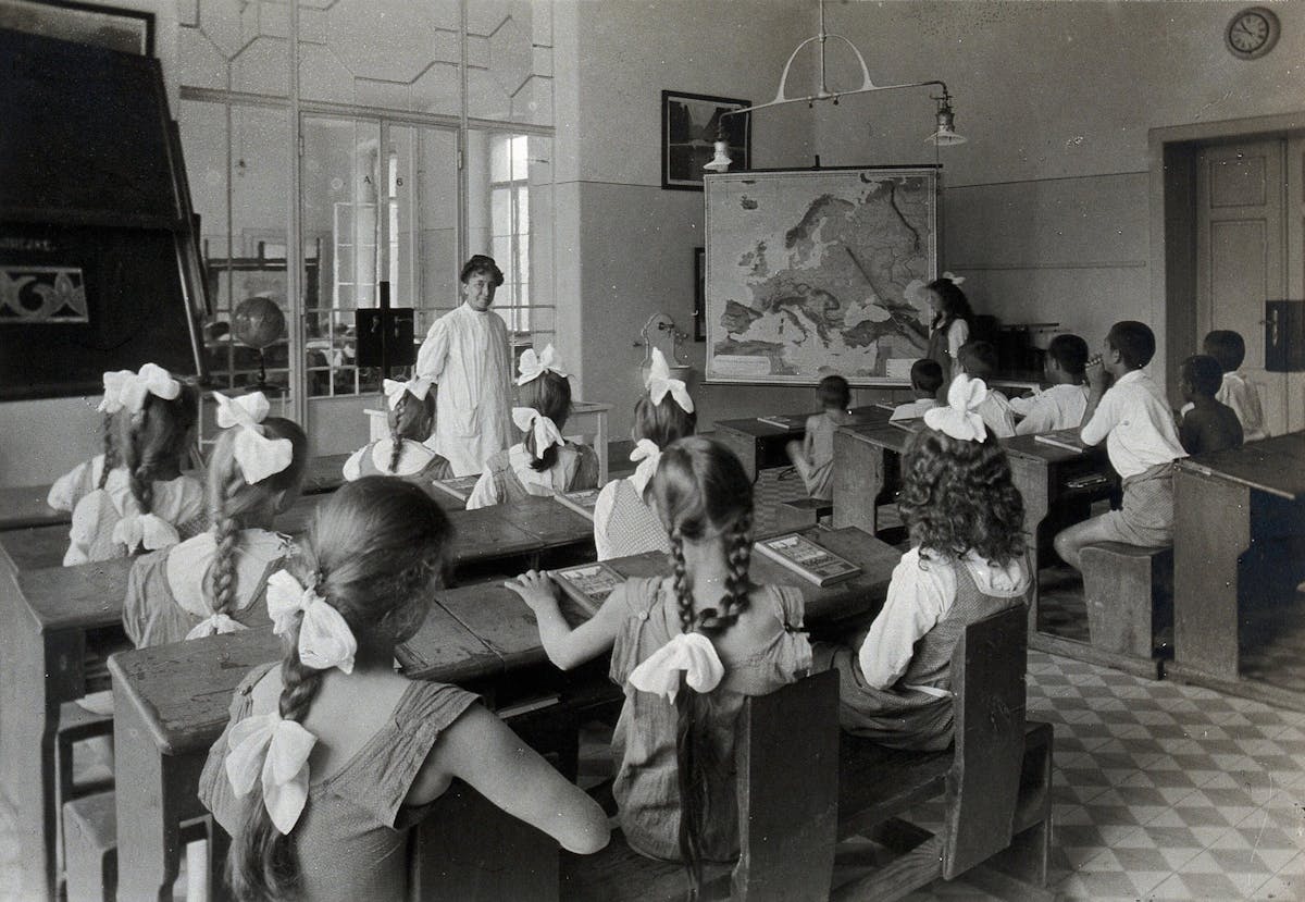 Black and white photograph of a classroom with a teacher in a white gown standing, looking at the children in front of her. Three rows wooden benches with tables attached have girls and boys seated, girls on one side, boys on the other, looking at a young girl pointing at a map of Europe with a stick next to the teacher.