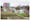 Photograph of an allotment site divided into different plots with grass pathways. In the background there is a large modern building made from red brick and glass to the top left of the image. In the bottom right there is a small rundown shed and a greenhouse, next to which is a small crop of bright yellow daffodils.