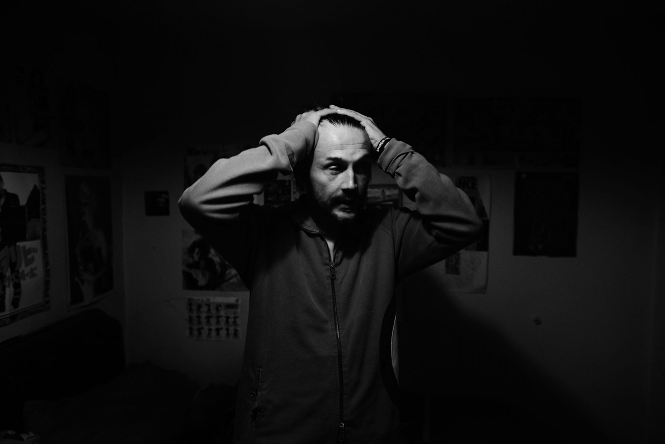 Black and white photograph showing a man standing in the middle of a dark room, his hands clasped to his head.