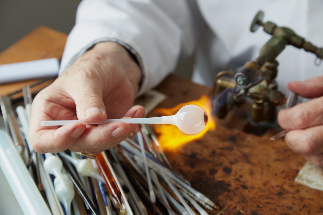 Photograph of the hands of a man in a white lab coat, holding a tube of white glass with a sphere resembling an eye ball at one end, to the flame of a gas torch. Next to him on the workbench are thin tools and more lengths of glass tubing.