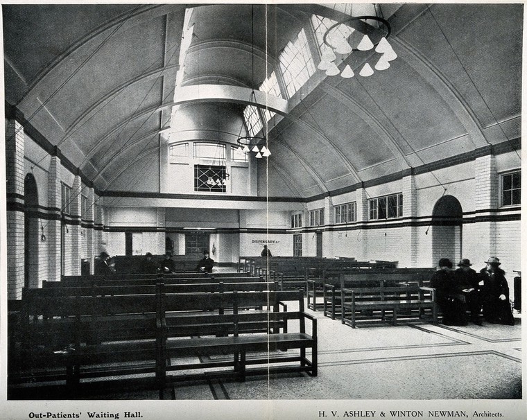 Black and white photograph of a waiting room. It is filled with rows of mostly empty wooden benches. A group of three people sit close together on one of the benches. Four people sit spaced further apart on another. A person sits alone at the very back of the room beneath a sign on the wall that reads: 'Dispensary'.