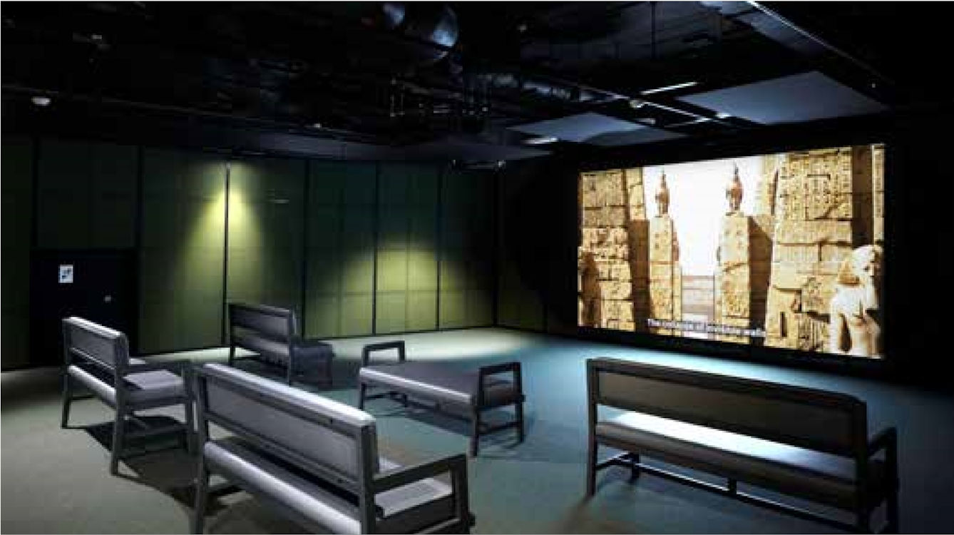 A film plays on a large screen taking up most of the wall in a darkened room. Five long benches are arranged in two rows facing the screen for viewing.