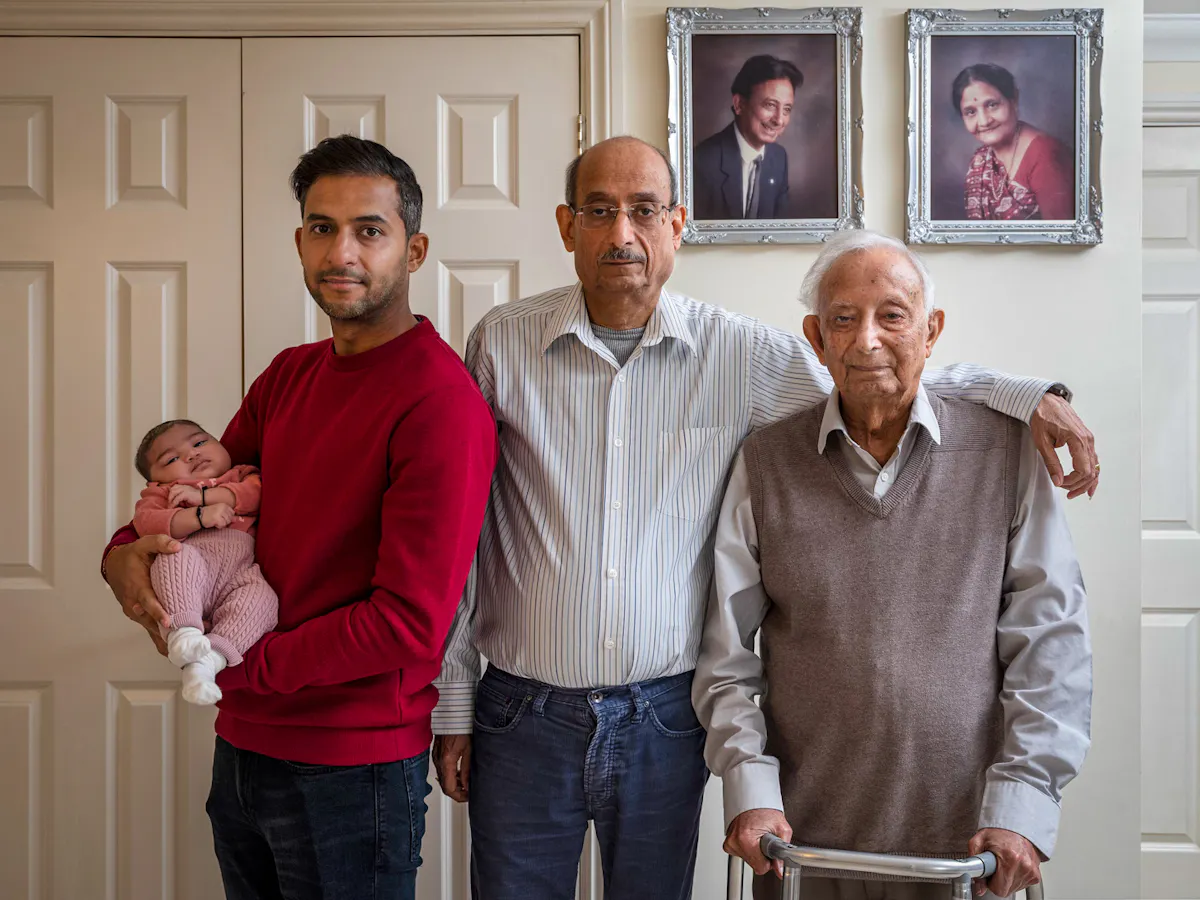 Family group photograph showing four generations of the same family. The great-grandfather stands on the right holding a walking frame, by his side is the grandfather whose arm rests on the great-grandfather's shoulder. Next to him is the grandson who cradles the great-granddaughter who is a baby in his arms. They are stood in front of a white wall on which two studio portraits are hung in silver frames, showing the great-grandfather and his wife many years before. They all look straight into the camera.
