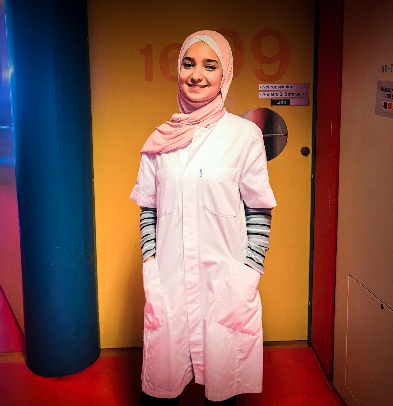 Photograph of a young woman wearing a head scarf and dressed in a medics coat. Behind her are the colourful doors and walls of a hospital.