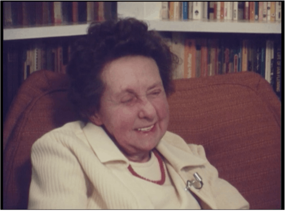 Film still of an older woman smiling. She's sat on a red armchair with bookshelves in the background.