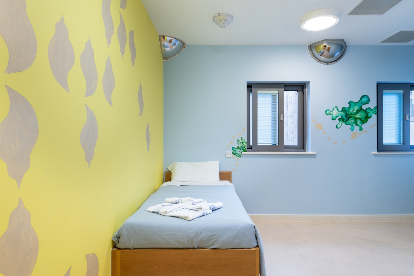 A photograph showing a room containing a single bed neatly made. The wall on the left-hand side of the image is painted lemon yellow with silver whelk shells of about 30 cm spaced at varying intervals on it. The adjacent wall is painted sky blue on which there are two simple images of green seaweed in two tones of green. Above the bed, in the top left corner there is a quadrant shaped 90 degree visibility mirror, and along the ceiling a meter or so there is an additional semi-circular 180 degree visibility mirror.