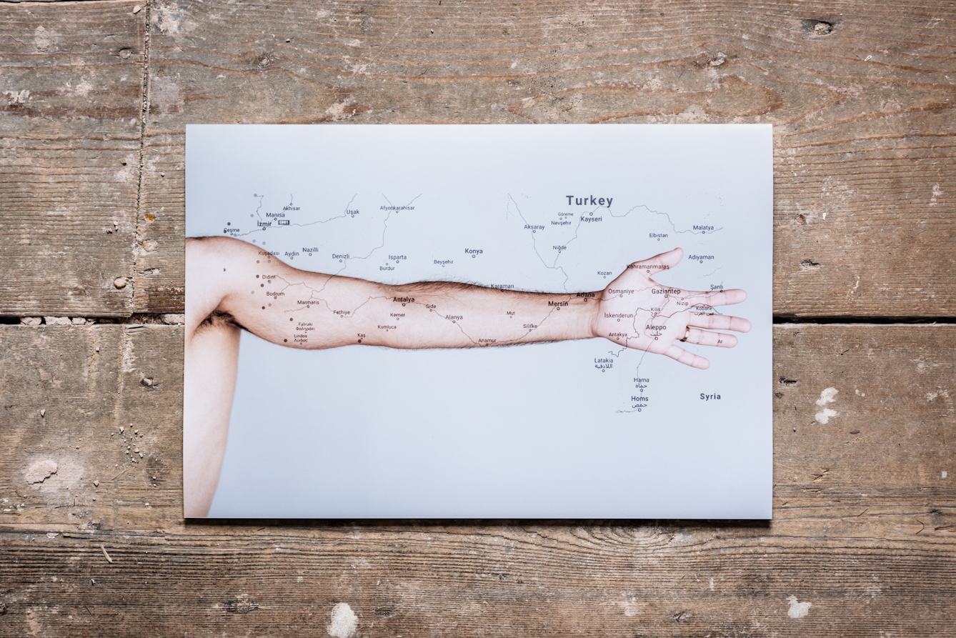 Photograph of a photographic print on a weathered, paint spattered wooden floor. The print shows a naked human arm outstretched against a white background. Superimposed on the arm is a simple line map in white, showing roadways and place names.
