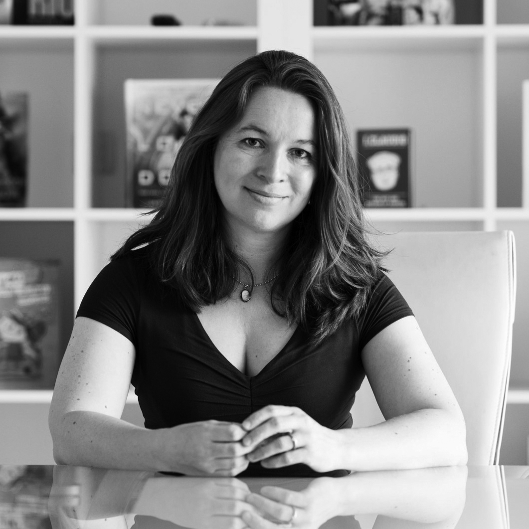 Photograph of the upper body of a woman with shoulder-length hair, sitting with her elbows on a table, in front of a set of shelves. 