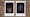 Photograph of two Instax Mini instant film prints in a line, resting on a textured brick surface. The two prints feature the same individual. The print on the left shows the individual emerging out of darkness with their left arm raised up up to show their armpit hair. The print on the right shows the individual's face also emerging out of darkness, spotlit. The blue background can be seen behind their head and the golden glint of one of their earrings.