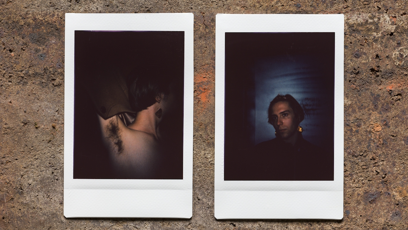 Photograph of two Instax Mini instant film prints in a line, resting on a textured brick surface. The two prints feature the same individual. The print on the left shows the individual emerging out of darkness with their left arm raised up up to show their armpit hair. The print on the right shows the individual's face also emerging out of darkness, spotlit. The blue background can be seen behind their head and the golden glint of one of their earrings. 