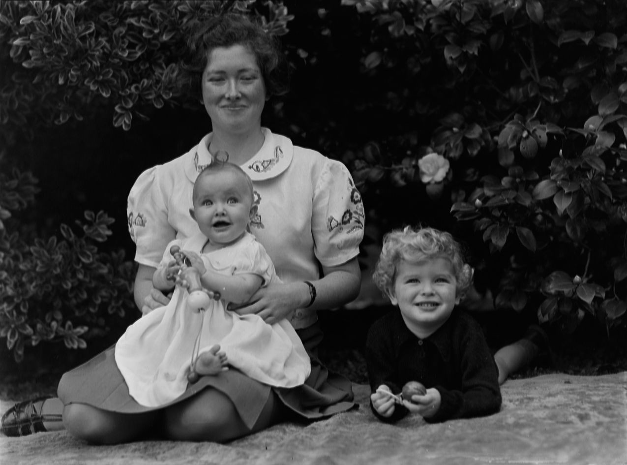 A young woman sitting with a baby and toddler. They are sitting outside on a rug in front of a bush. The woman is wearing an embroidered blouse and has her legs crossed under her. She holds the baby on her lap and her eyes are partially closed. The baby and the toddler both hold toys.