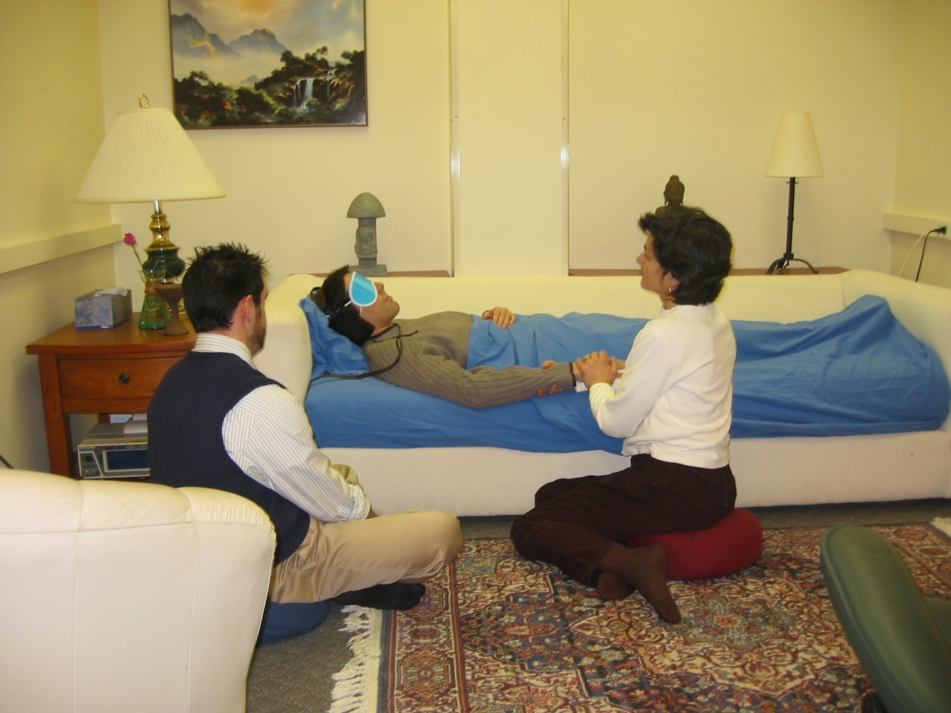 A photo of one of the comfortable rooms at Johns Hopkins University used in clinical studies to determine the effects of psilocybin and other hallucinogenic drugs. Two guides are seated beside a figure lying on a sofa.