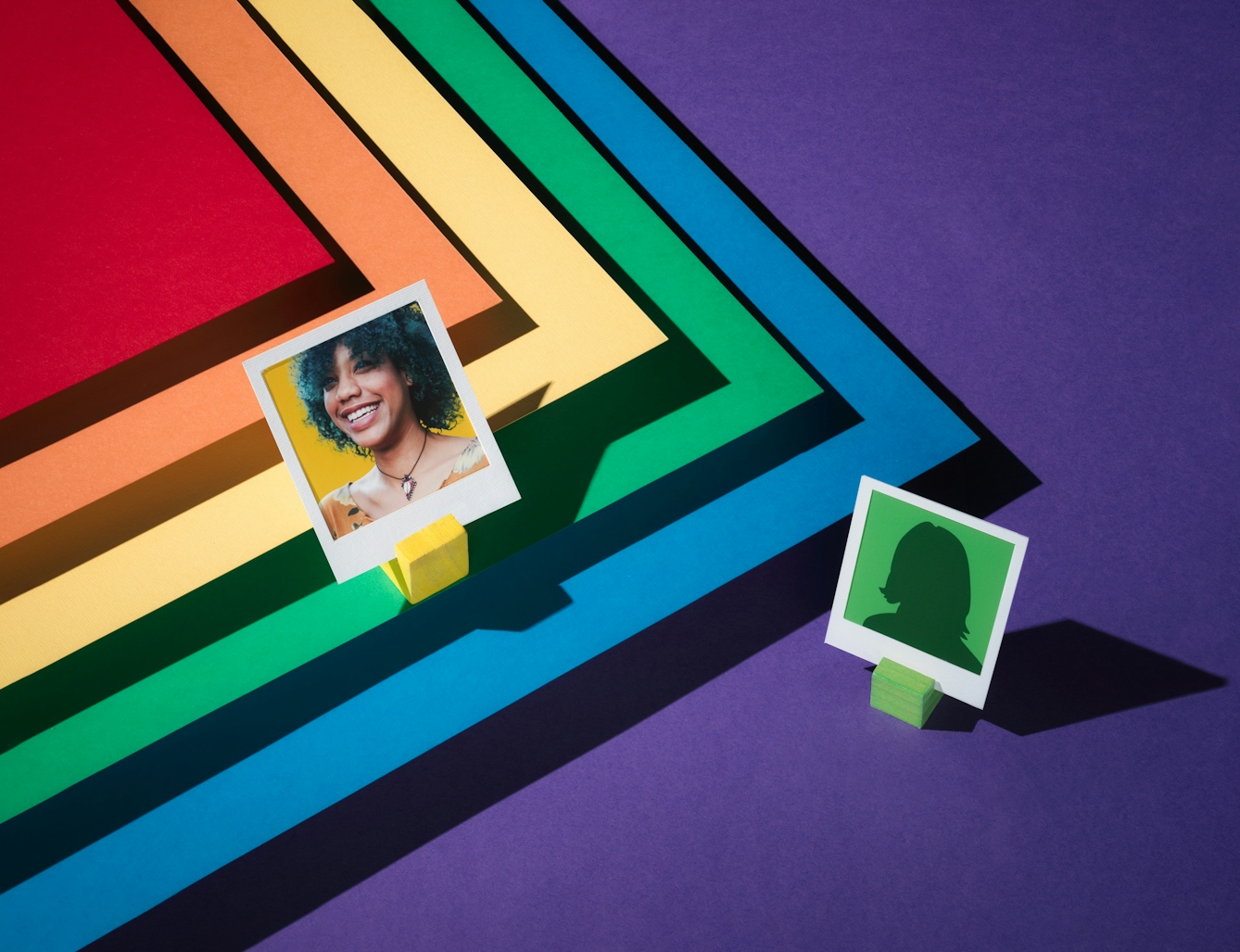 Photograph of a tiered set made up of the LGBTQ+ colours of red, orange, yellow, green, blue and purple.  The tiers form a chevron.  On the second tier, the green one, the portrait of a woman against a yellow background is framed in a small white frame.  Below this image, to the right, an anonymous figure on a green background is facing towards her.