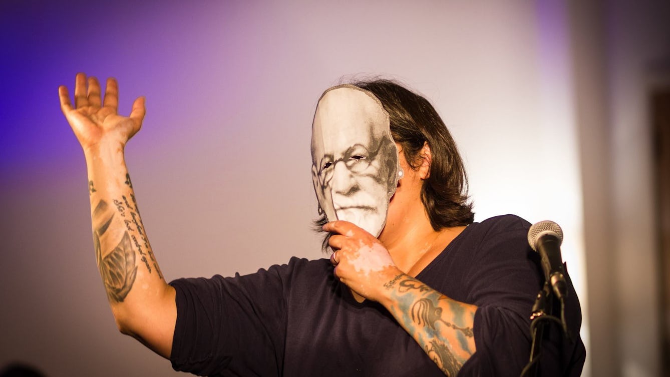 A woman with shoulder-length hair and tattooed arms holds a black and white mask of Freud's face over her own, and gesticulates. 